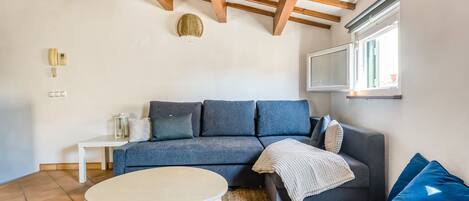 A living room with a sofa, a round table, a patterned rug, wooden beams on the ceiling, and a window with blinds.