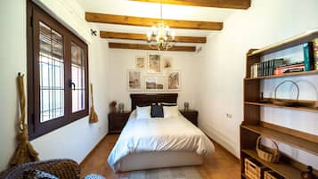 Ein Schlafzimmer mit Bett, Holzbalken, einer Chandelieber, einem Bücherregal und einem Fenster mit Jalousien.