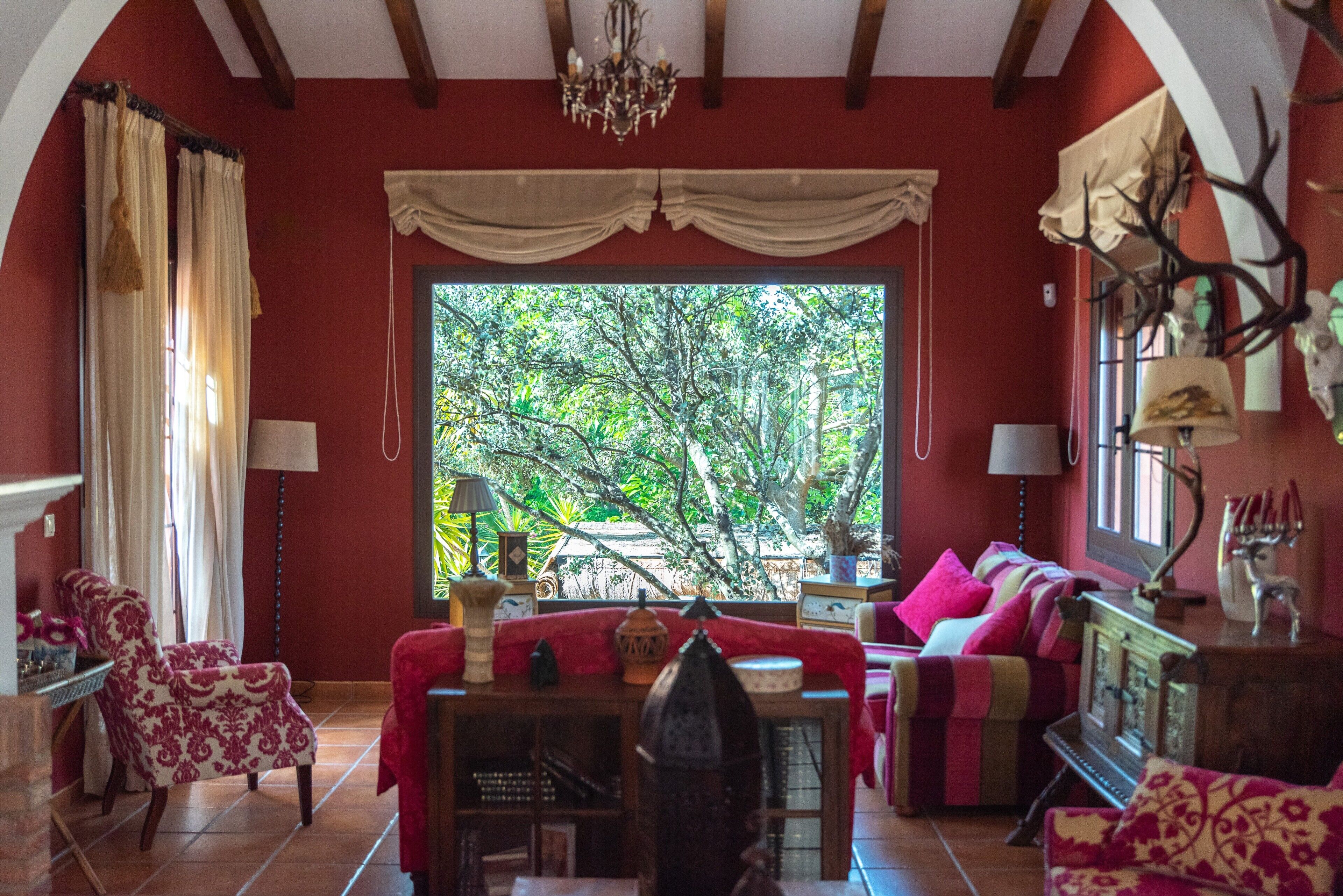 A room with a red wall, a large window with a view of trees, a chandelier, and a sofa with a red cover.