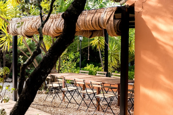 A covered outdoor seating area with a wooden table and chairs, surrounded by greenery.