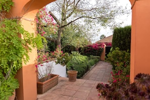 A pathway flanked by potted plants and flowering shrubs, leading to a building with a tiled roof.