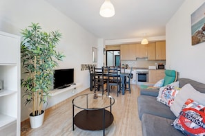 A modern living room with a sofa, a glass coffee table, a TV, a dining area with chairs, a kitchen in the background, and a potted plant. - Casa 'Tazacorte' with Sea Views, Shared Pool and Wi-Fi (Tazacorte)