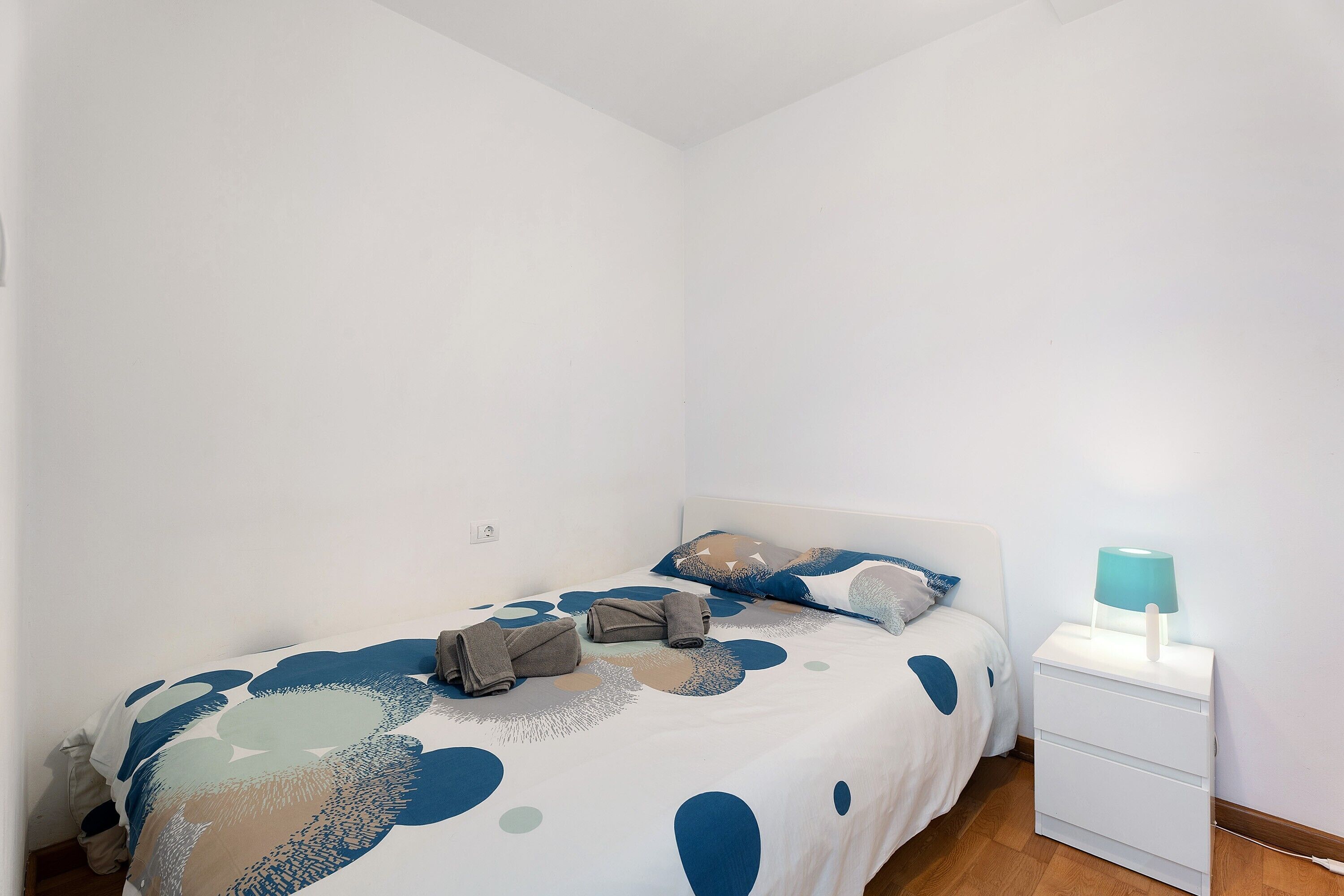 Quarto pequeno e limpo, com uma cama, mesa de cabeceira com abajur e uma tomada na parede.