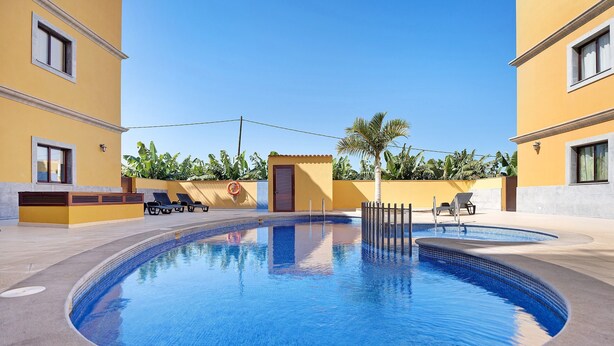 Una piscina con lettini, una palma e un edificio giallo.