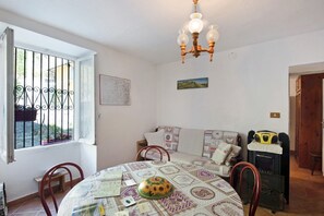 Un coin repas avec une table ronde, des chaises, un lustre, une fenêtre avec des barreaux, un canapé, une radio et un petit banc.