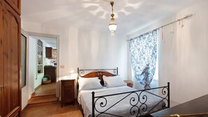 Une chambre à coucher avec un lit en métal, une table de chevet, une commode, un lustre, une fenêtre avec des rideaux à motifs et une armoire en bois.