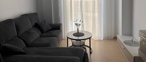 Een moderne woonkamer met een donkere bank, een kleine ronde tafel en een groot raam met transparante gordijnen.