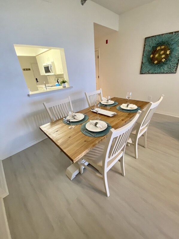 A dining area with a wooden table set for four, white chairs, and a kitchen visible through an open door. - Oceanfront Condo, Private Balcony (Palm Coast)