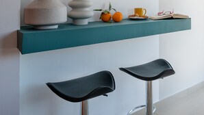 A teal shelf with two black stools, a vase, a decorative object, oranges, a yellow mug, and a book with glasses on top. - Meros Filikon - Stylish Two Bedroom apt. (100m2) (Zakinthos)