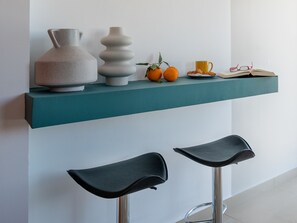 Un estante azul verdoso con dos taburetes negros, un florero, un objeto decorativo, naranjas, una taza amarilla y un libro con gafas encima.
