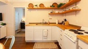 Une cuisine de taille réduite avec des meubles blancs, un plan de travail en bois et une cuisinière. Des étagères en bois sont fixées au-dessus du plan de travail et servent à ranger les ustensiles de cuisine.