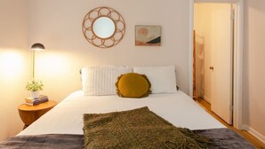 Un lit bien fait, avec une literie blanche et un oreiller de couleur moutarde, une petite table de chevet avec une lampe et des livres, un miroir fixé au mur et une porte donnant sur une autre pièce.