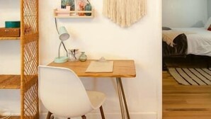Una habitación con un escritorio de madera, una silla blanca, un tapiz de macramé y un estante de mimbre.