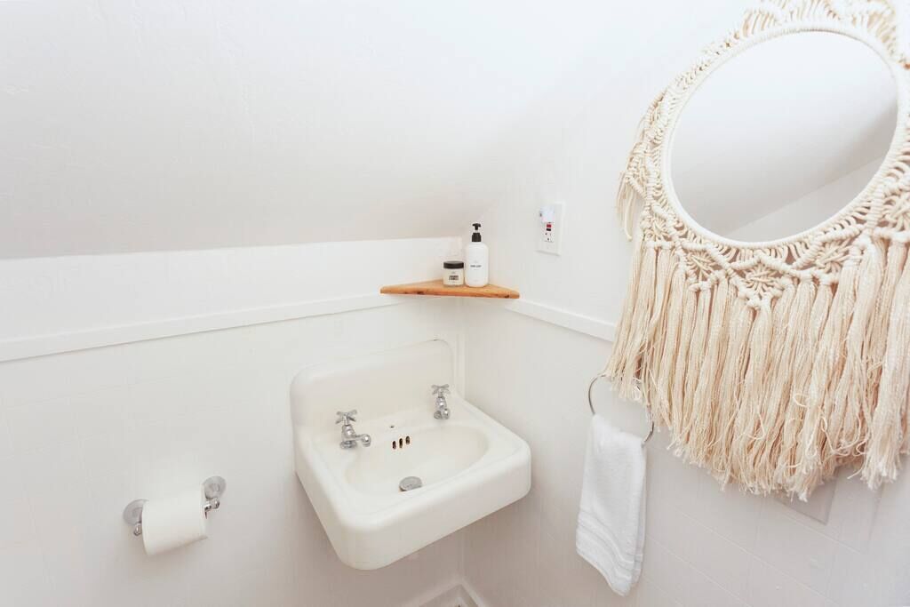 A bathroom with a white sink, a mirror with macrame fringe, and a wooden shelf with toiletries.