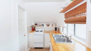 Eine kleine Küche mit weißen Schränken, einer Holz Arbeitsplatte, einem eingebauten Herd und einem Fenster mit einer Bambus Jalousie.