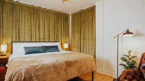 Un lit bien fait, recouvert d’une couverture beige, avec deux oreillers et une lampe de chevet. La pièce est dotée d’un plancher en bois et de rideaux verts.