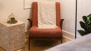 Habitación con televisor de pantalla plana montado en la pared, lámpara de pie, sillón rojo con una manta blanca, mesita de noche con estampado y una planta pequeña.