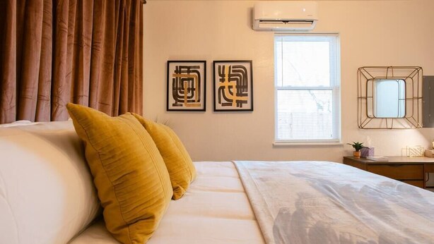 Una habitación de hotel con cama, ventana con persianas, aire acondicionado y dos cuadros enmarcados en la pared.