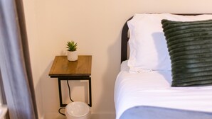 Un lit avec une literie blanche et un oreiller vert, une petite table de chevet en bois avec un pot de plante, et une poubelle blanche.