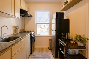 Cocina compacta con lavabo, estufa, horno y refrigerador. Hay una ventana con persianas, un cubo de basura pequeño y una planta en maceta sobre la encimera.