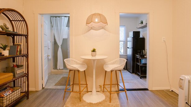 Un espace de vie compact comprenant un coin repas avec deux chaises blanches et une table ronde, une bibliothèque en bois et une cuisine visible à travers une porte ouverte.