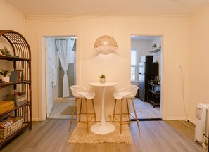 Un espacio habitable compacto con comedor, que incluye dos sillas blancas y una mesa redonda, una estantería de madera, y una cocina visible a través de un arco.