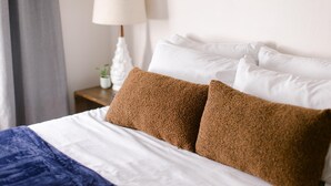 Una cama bien hecha con almohadas blancas y marrones, una lámpara blanca en una mesita de noche y una cortina gris.