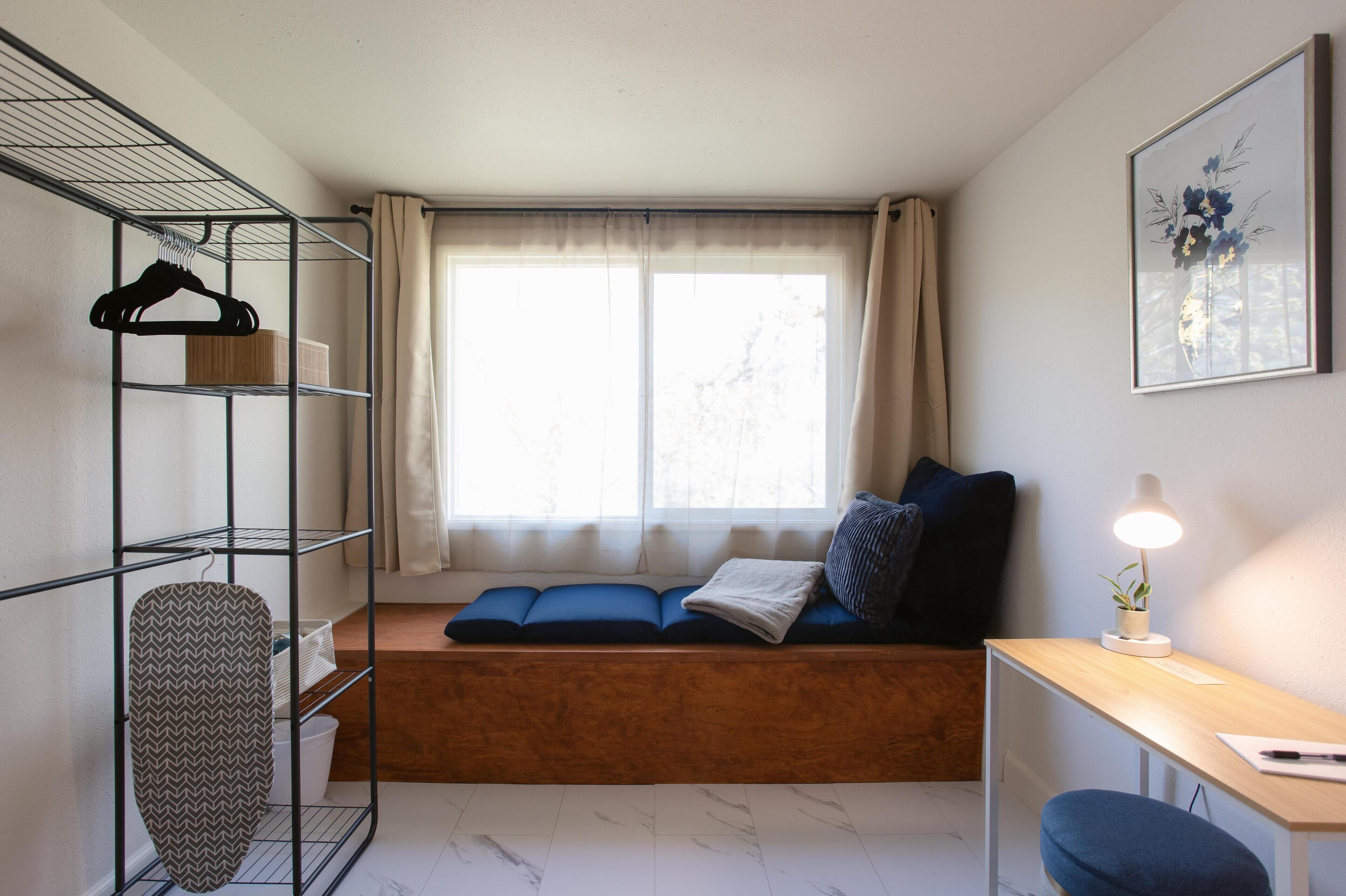 A compact room with a wooden bench, a desk, and a shelving unit with hangers and an ironing board.