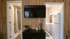Un pasillo con un televisor montado en la pared, paneles de madera, un mueble bajo con una planta y una cocina visible a través de una puerta abierta.