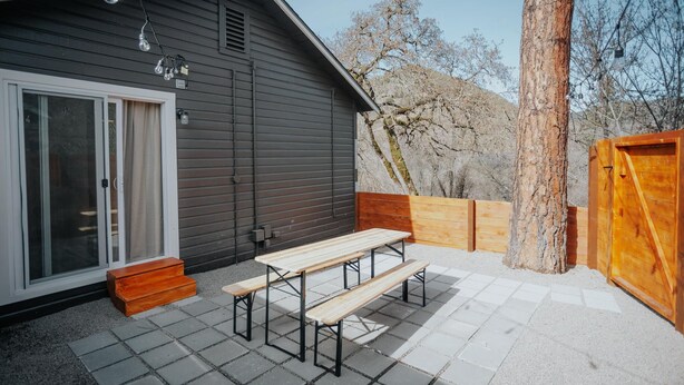 A patio with a wooden table and benches, a sliding glass door, and a wooden fence. - Located in a scenic area perfect for travelers along I-5. You are right by the Rogue River, offering stunning views and tranquility, yet conveniently positioned for easy travel access. (Gold Hill)