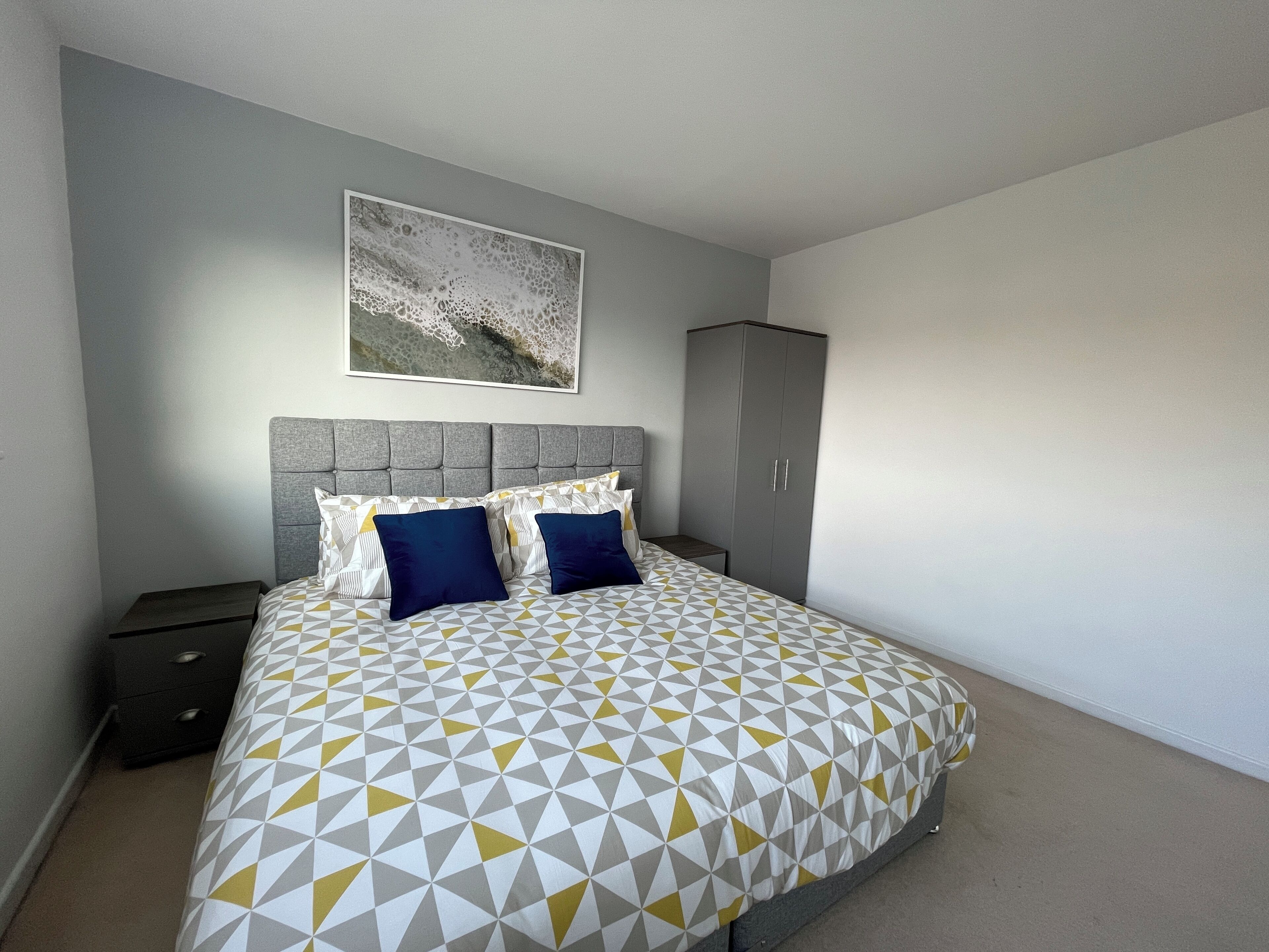 A bedroom with a bed, a grey headboard, a grey wardrobe, and a wall-mounted artwork.