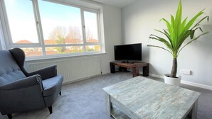 A living room with a grey armchair, a wooden coffee table, a television on a stand, and a potted plant. - Bright & Airy 3BR Home with Parking (Sheffield)