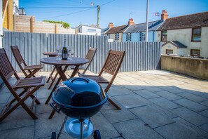 A patio with a round table, four wooden chairs, and a barbecue grill. - Ivy Cellar - 1 Bedroom Apartment - Tenby (Tenby)
