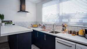 A modern kitchen with a central island, dark cabinets, a built-in dishwasher, and a window with blinds. - Ivy Cellar - 1 Bedroom Apartment - Tenby (Tenby)