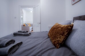 A neatly made bed with grey linens and a brown pillow, a white door leading to another room, and a radiator on the wall. - Ivy Cellar - 1 Bedroom Apartment - Tenby (Tenby)