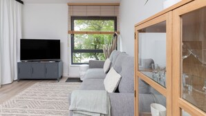A modern living room with a grey sofa, a television on a grey cabinet, a large window with blinds, and a plant on a wooden shelf.