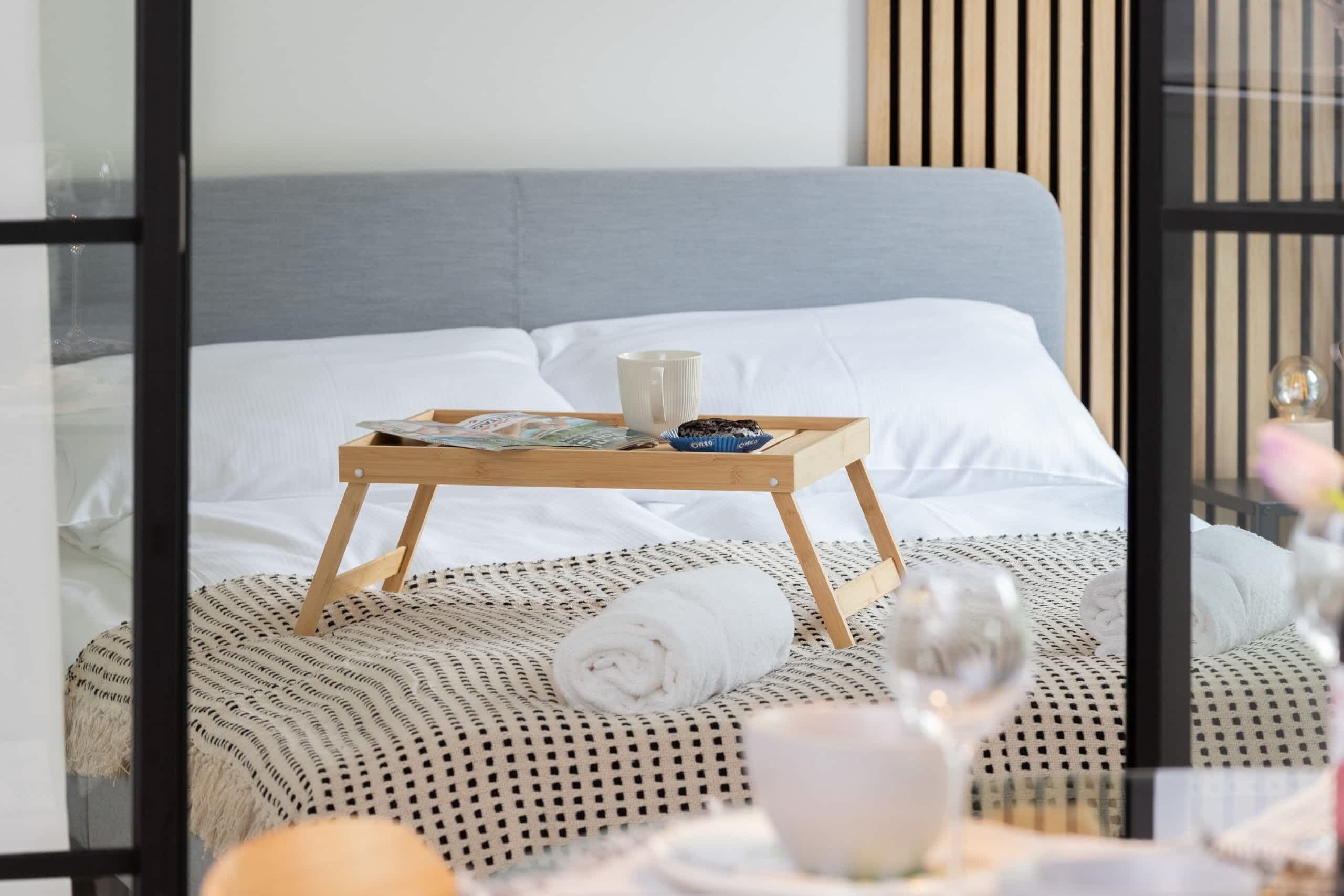 Een net opgemaakt bed met een witte handdoek, een dienblad met een kopje en een muffin, en een glas water op een tafel.