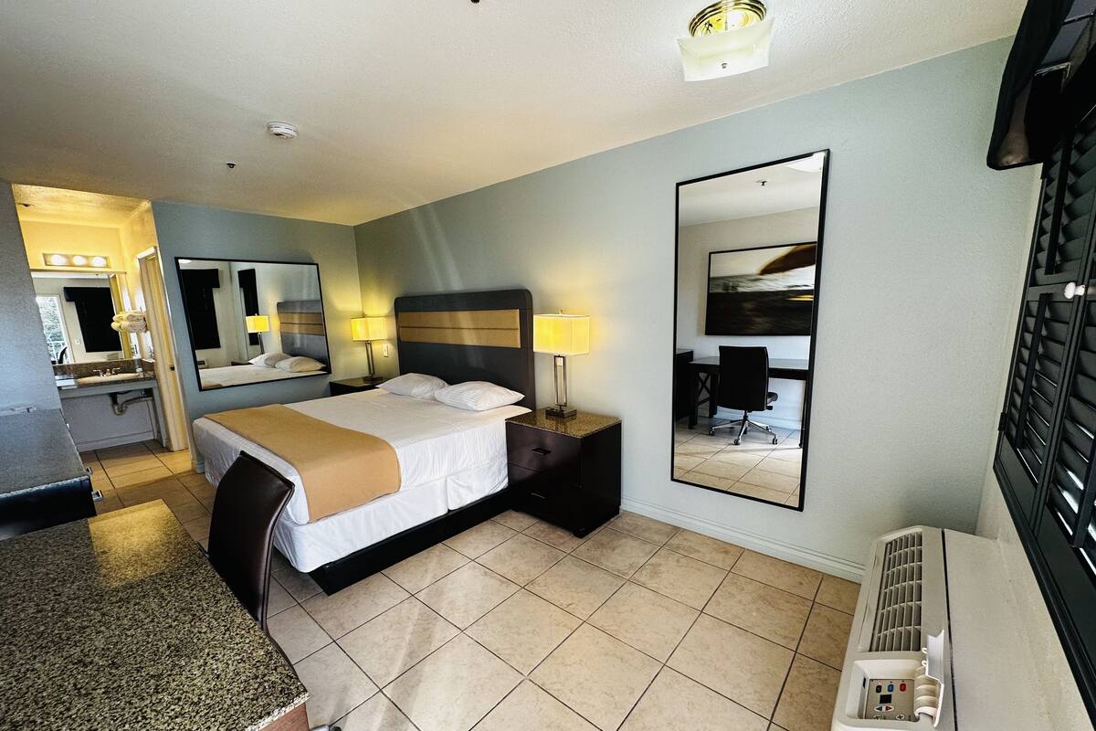 Ein Hotelzimmer mit Bett, Nachttischlampen, einem großen Spiegel, einem Schreibtisch und einem durchgegangenen Tür sichtbar erkennbaren Badezimmer.