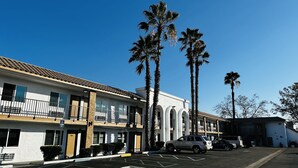 A parking lot with a white building featuring a tiled roof and a white facade. There are palm trees in front of the building. - Casa Blanca Express & Suites Cypress Buena Park - Anaheim Area (Cypress)