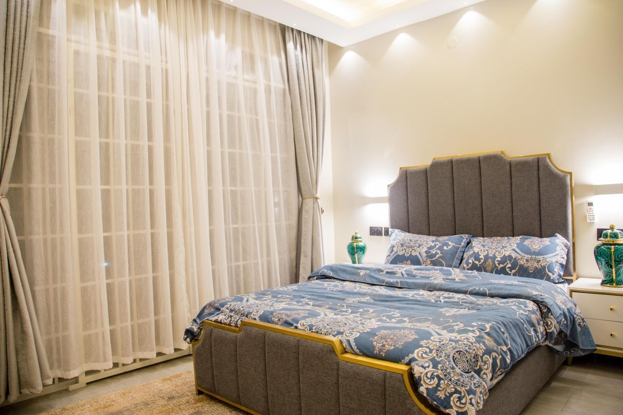 Une chambre à coucher avec un grand lit, une table de chevet, une lampe et une fenêtre ornée de rideaux légers.