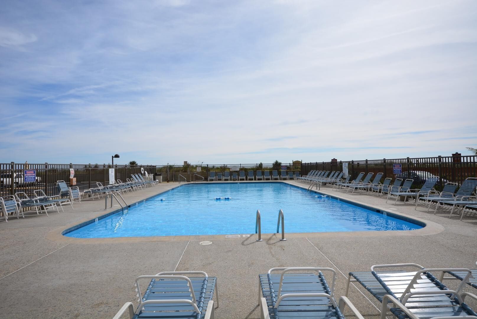 Ein rechteckiger Swimmingpool mit gefliestem Sonnendeck, umgeben von Liegestühlen und einem Metallzaun.