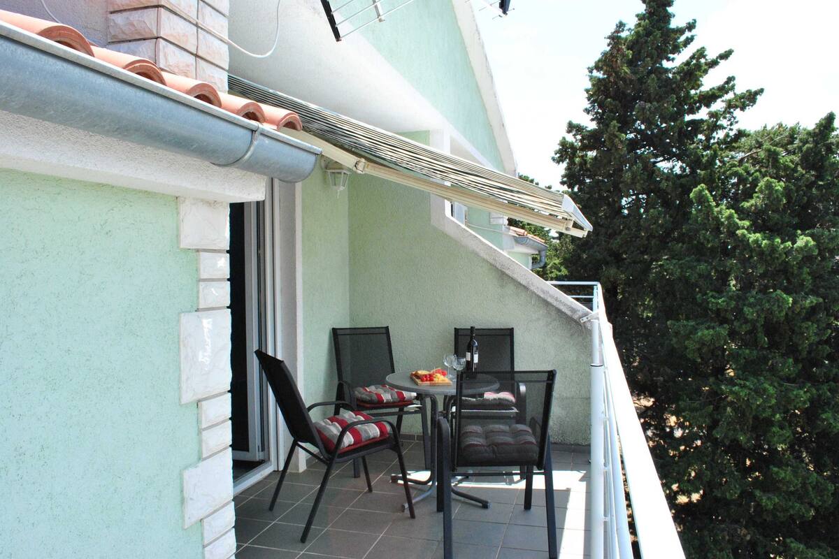 A balcony with a table and chairs, a window, and a tree in the background.