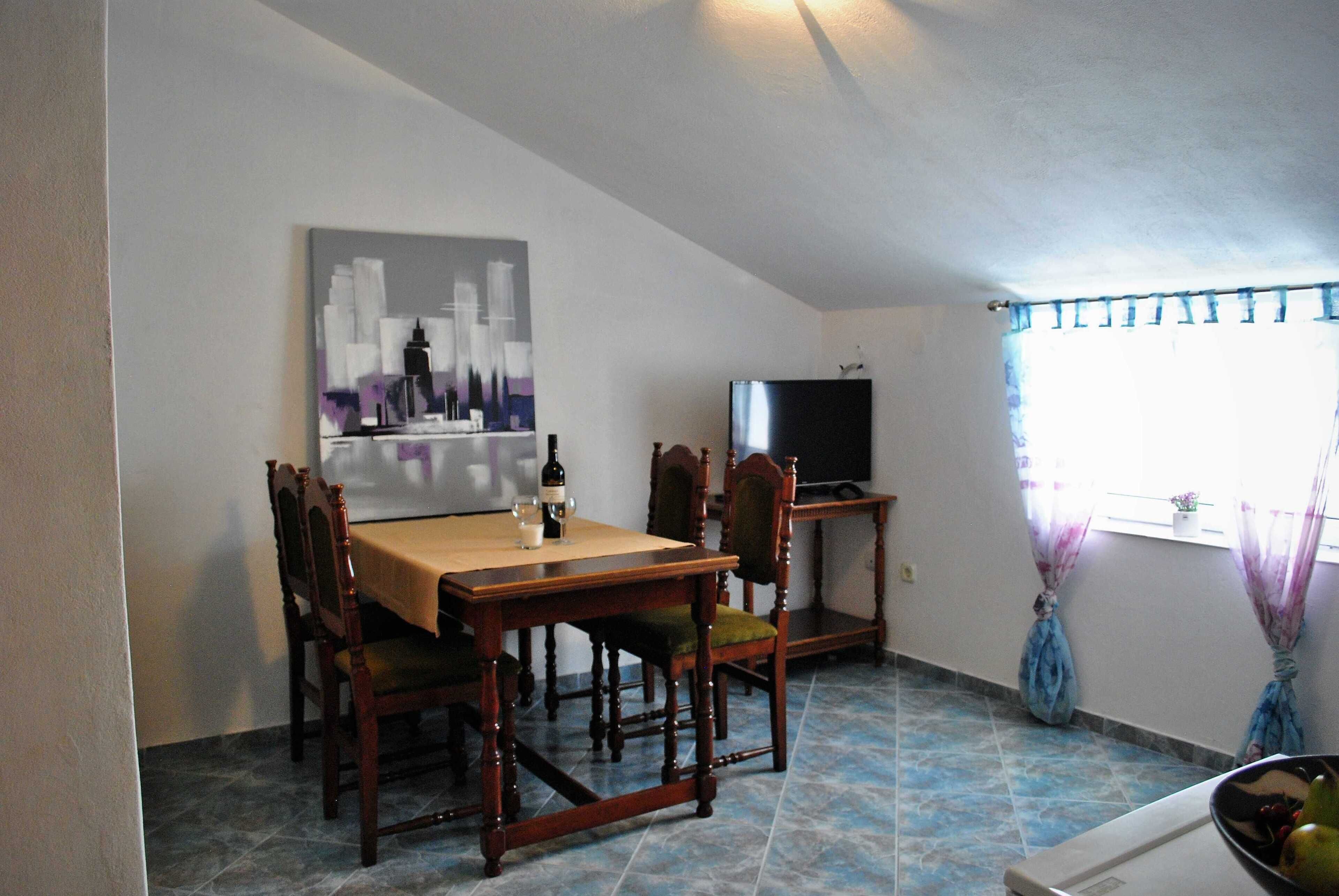 A dining area with a table and chairs, a painting on the wall, a television on a stand, and a window with curtains.