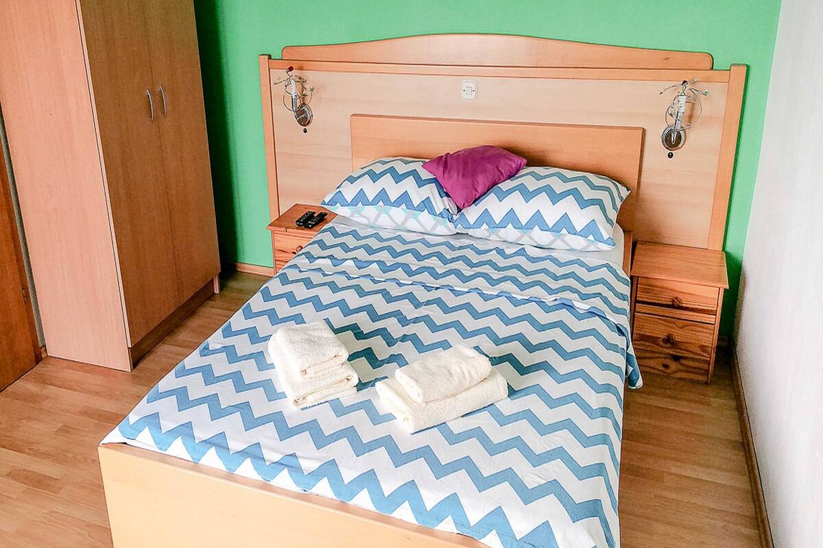 A single bed with a blue and white zigzag patterned bedspread, a wooden headboard, a nightstand with a lamp, and a wooden wardrobe.
