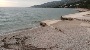 Uma praia de seixos com um molhe rochoso e montanhas ao fundo.