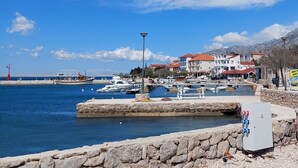 Uma marina costeira com barcos atracados, um muro de pedra em primeiro plano e edifícios com telhados vermelhos ao fundo.