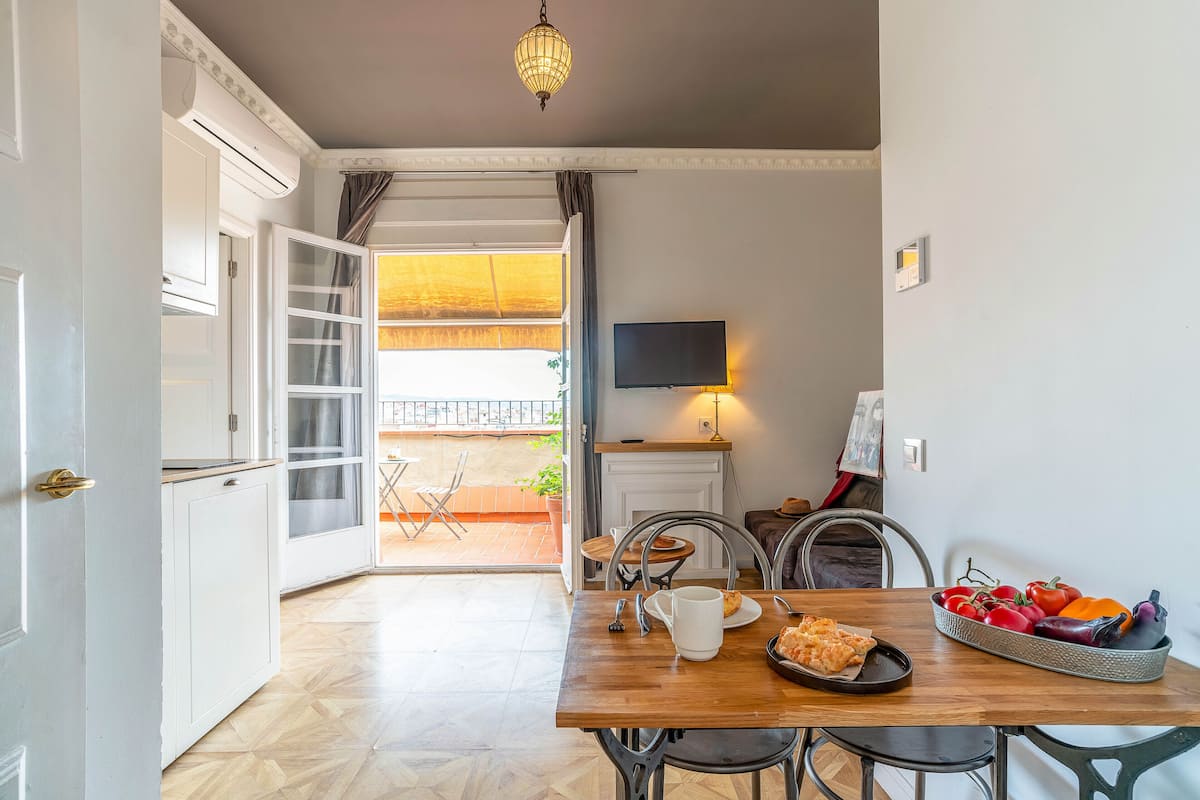 A dining area with a wooden table, chairs, a TV mounted on the wall, and a view of the outside balcony.
