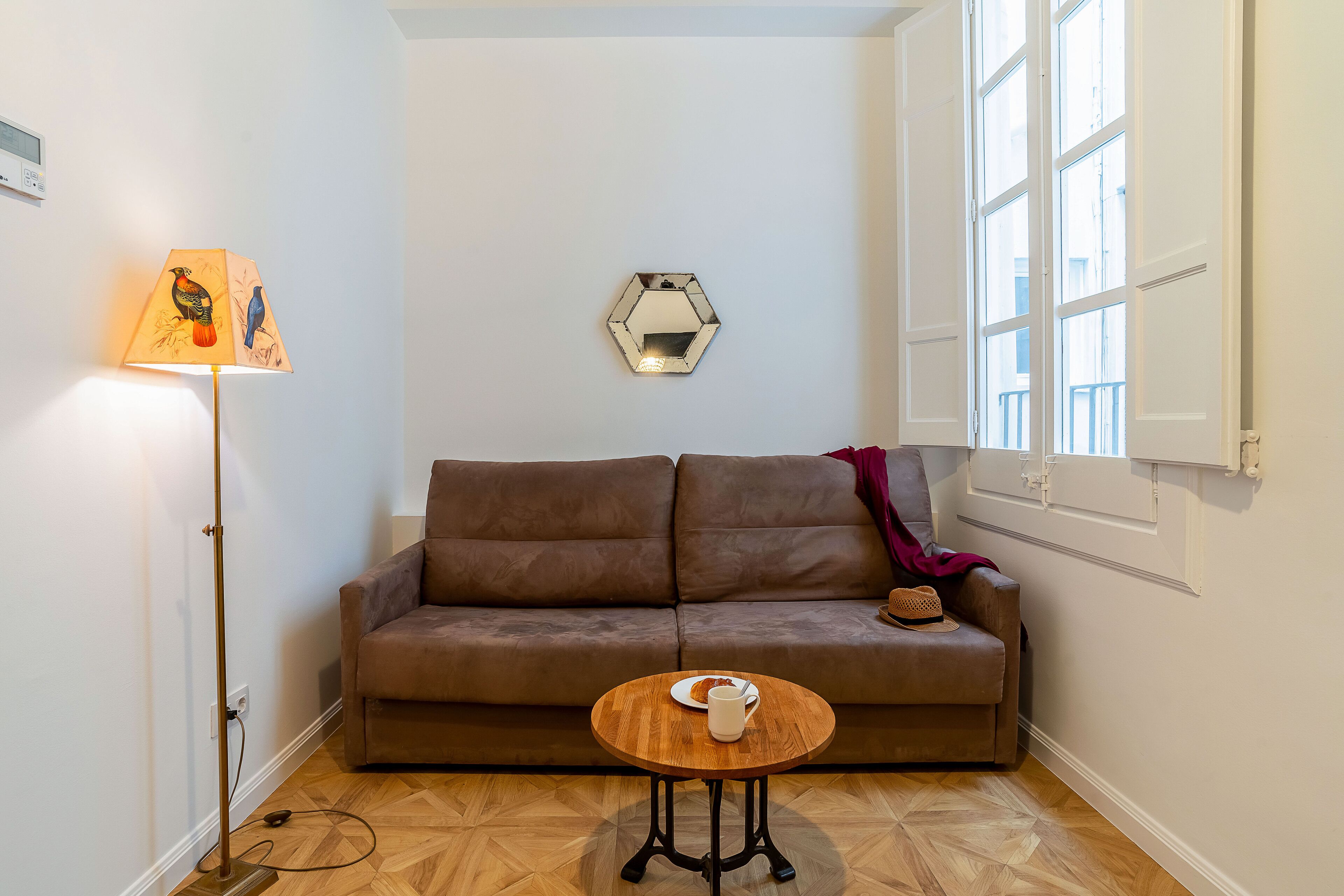 A cozy room with a brown sofa, a round wooden table with a white mug, a floor lamp with a bird design, and a hexagonal mirror on the wall.