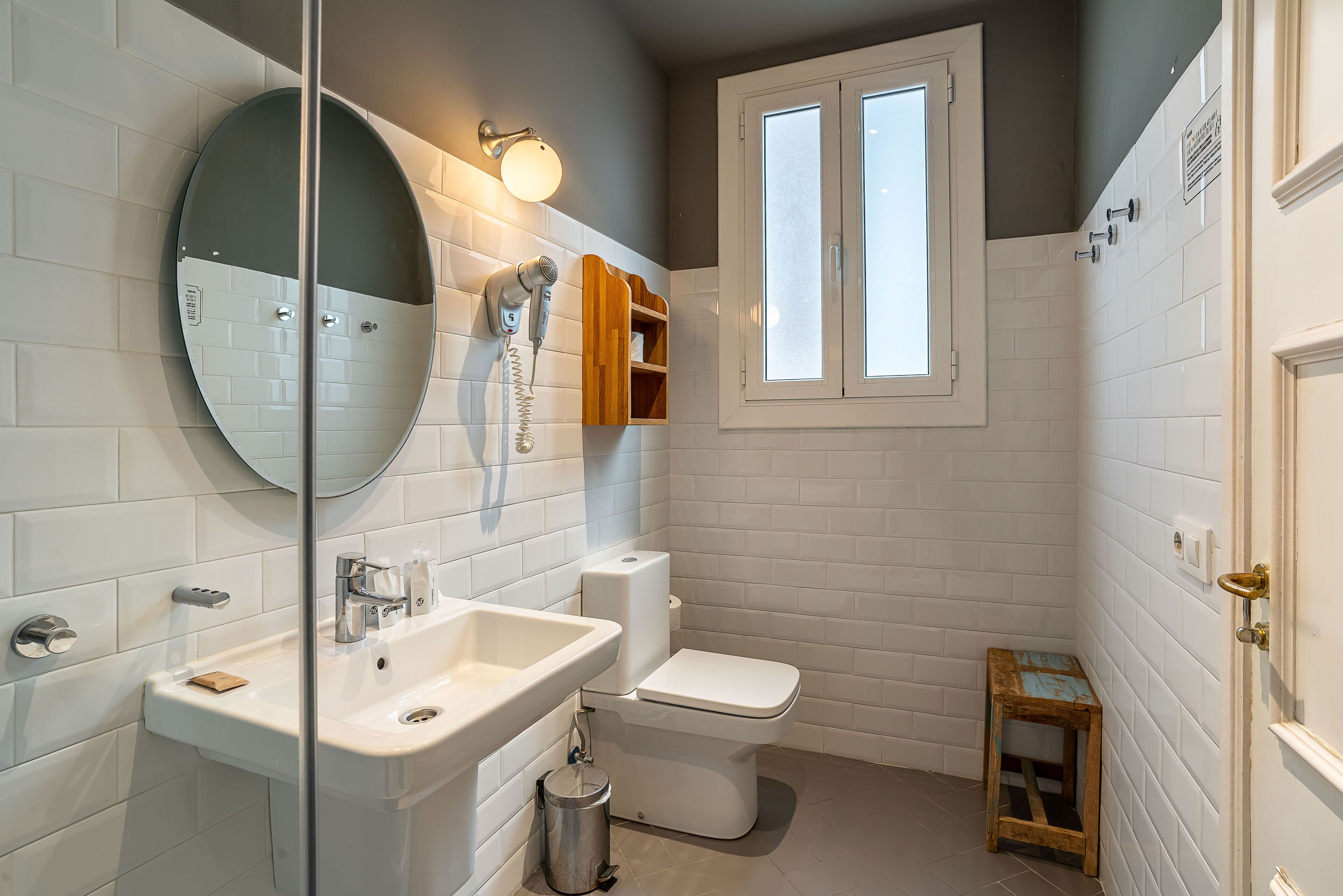 Ein Badezimmer mit weißem Waschbecken, Spiegel und Toilette, einem kleinen Holzschemel und einem Fenster über der Toilette.