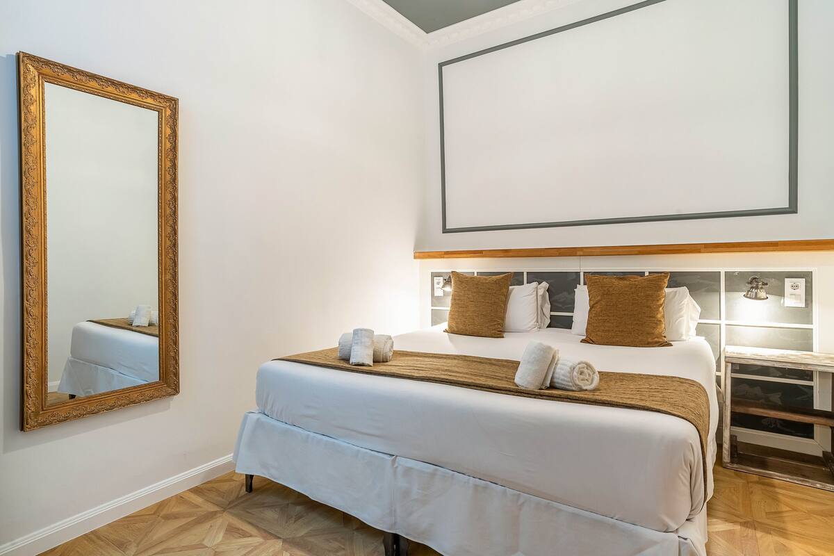 A hotel room with a large bed, a wooden headboard, a mirror, and a bedside table with a lamp.